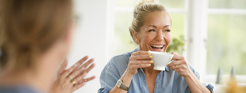 Zwei Frauen sitzen am Tisch, die eine Frau trinkt aus einer Tasse und lacht. 