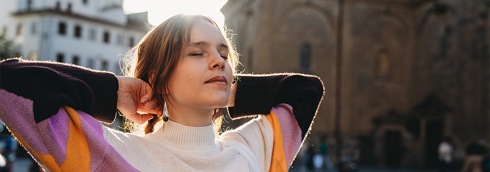Eine Frau steht in einer Stadt mit geschlossenen Augen und hält die Arme hinten an den Kopf.