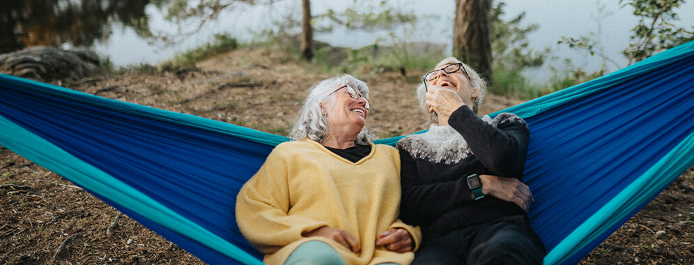 Zwei ältere Frauen liegen in einer Hängematte und lachen. 