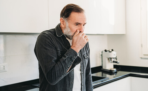 Ein Mann steht hustend in der Küche.