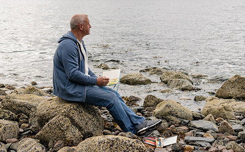 Ein Mann schreibt in ein Notizbuch, er sitzt am Meer.