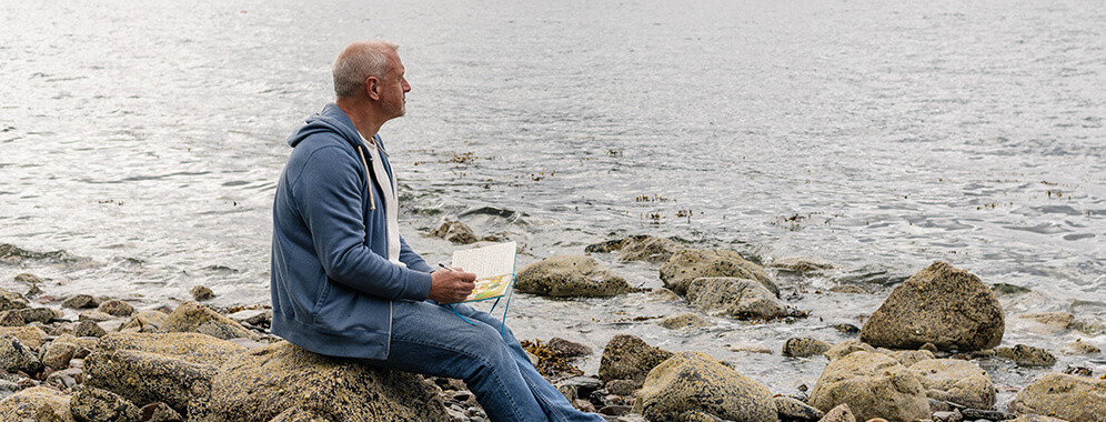 Ein Mann sitzt am Meer und schreibt in ein Notizbuch. 