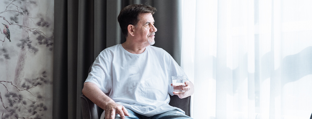 Ein älterer Herr sitzt auf einem Stuhl und trinkt ein Glas Wasser.