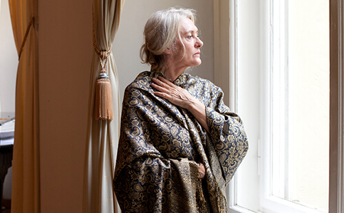 Eine Frau steht am Fenster und hält sich den Hals. 