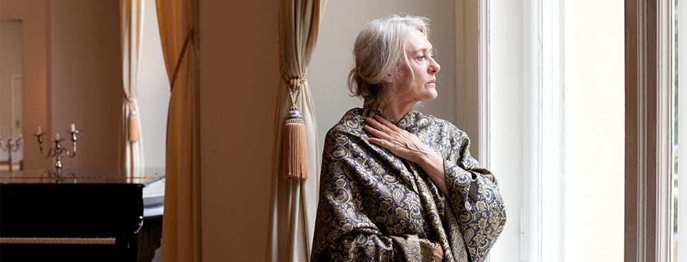 Eine ältere Dame steht am Fenster und hält sich den Hals. 