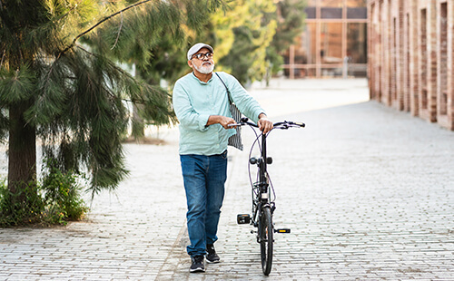Ein älterer Mann spaziert durch eine Stadt und schiebt dabei ein Fahrrad.