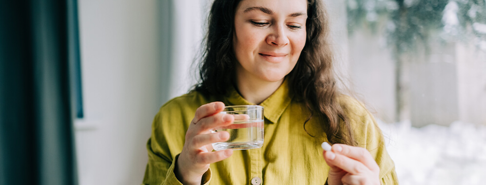 Eine Frau hält eine Abnehmpille in der einen und ein Glas Wasser in der anderen Hand und lächelt sanft.