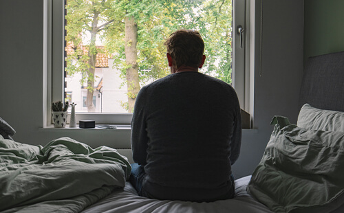 Man sieht einen Mann von hinten, der etwas zusammengesunken auf dem Bett sitzt und aus dem Fenster blickt. 