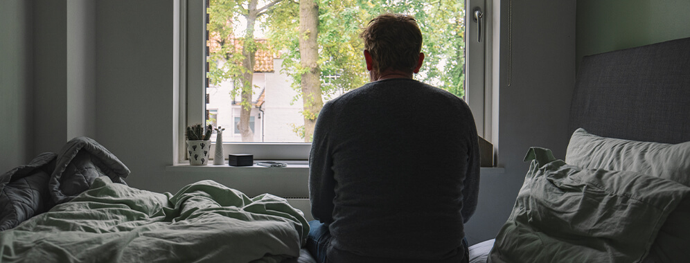 Man sieht einen Mann von hinten, der etwas zusammengesunken auf dem Bett sitzt und aus dem Fenster blickt.