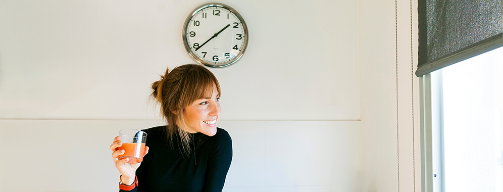 Eine Frau sitzt am Tisch, hinter ihr hängt eine Uhr.