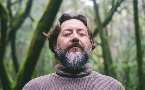 Ein Mann steht mit geschlossenen Augen in einem Wald und scheint tief zu atmen. 