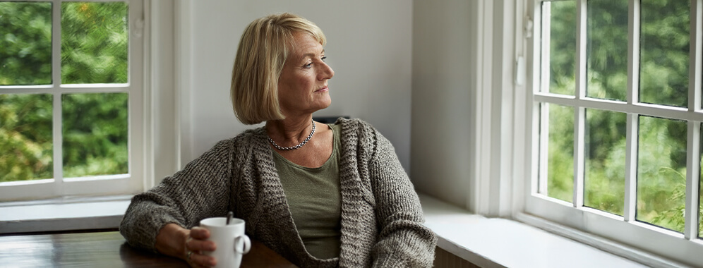 Eine Frau sitzt mit einer Tasse am Tisch und schaut aus dem Fenster. 