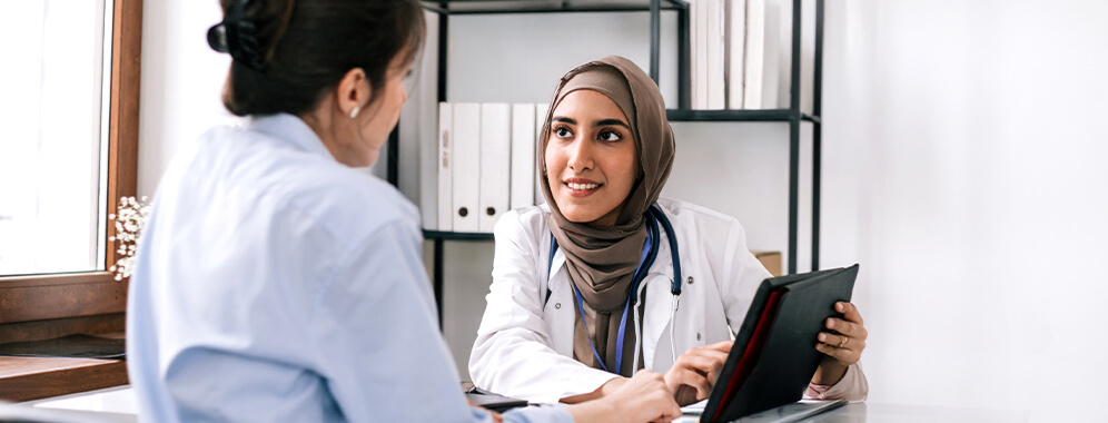 Eine Frau spricht in einer Praxis mit der Ärztin, die etwas auf dem Tablet zeigt.