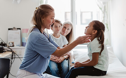 Ein Mädchen wird beim Kinderarzt untersucht, dahinter sitzen ihre Mutter und ihr Bruder.