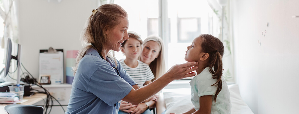 Ein Mädchen wird von einer Kinderärztin untersucht, im Hintergrund sitzen ihre Mutter und ihr Bruder.