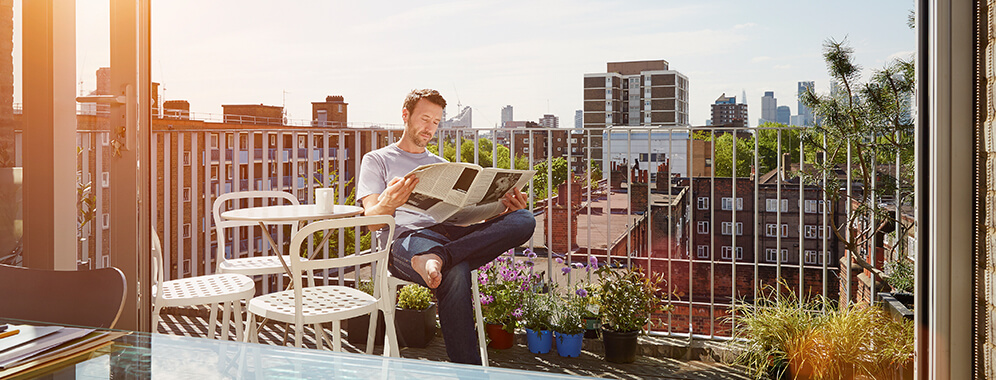 Ein Mann sitzt auf einem Balkon in der Sonne und liest Zeitung. 
