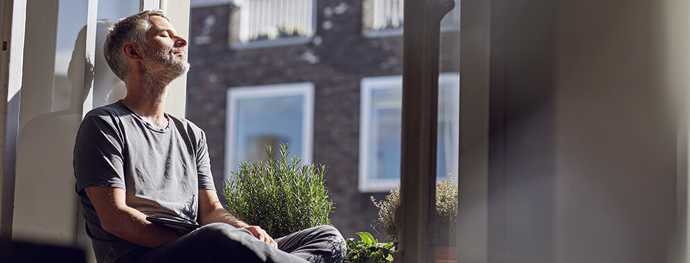 Ein Mann sitzt am offenen Fenster mit geschlossenen Augen und genießt die Sonnenstrahlen. 