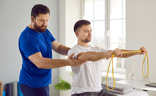 Ein Mann macht eine Übung mit einem Theraband, ein Physiotherapeut beaufsichtigt die Übung. 