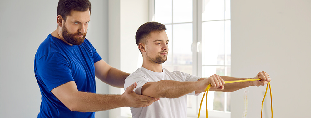 Ein Mann hält ein Theraband zwischen den Händen. Ein Physiotherapeut beaufsichtigt die Übung.