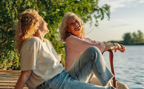 Zwei Frauen sitzen am See und lachen sich an.