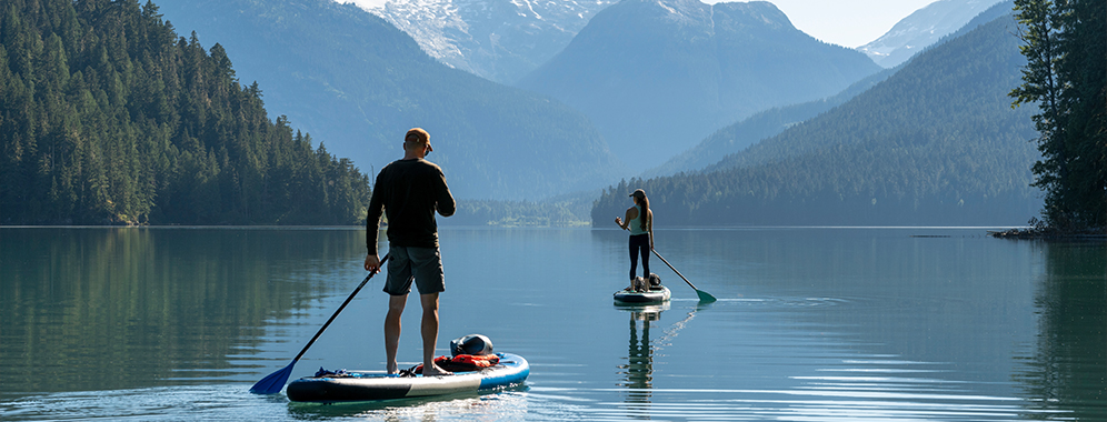 Eine Frau mit MS fährt mit einem Standup-Paddle-Board über einen See. Ein Mann begleitet sie auf einem eigenen SUP. 