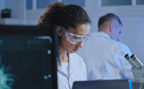 Eine Frau arbeitet im Labor, sie trägt eine Laborbrille und man sieht einen Bildschirm und ein Mikroskop.