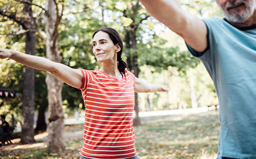 Eine Frau mit MS macht Sport im Park, ihre Arme sind zu den Seiten ausgestreckt. 