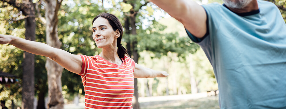 Frau mit MS macht Sport im Park, sie streckt die Arme zu den Seiten aus. 
