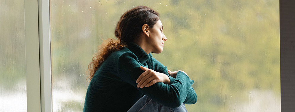 Junge Frau sitzt vor Glasfront und schaut nachdenklich hinaus.