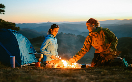 Zwei Frauen sitzen im Dämmerlicht an einem Feuer, im Hintergrund sieht man ein aufgebautes Zelt. 