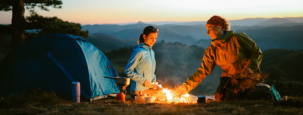 Zwei Frauen sitzen am Feuer, daneben sieht man ein Zelt. 