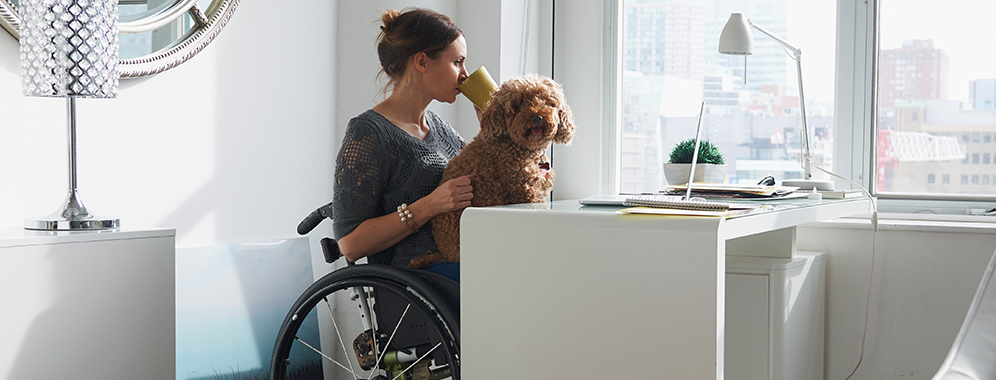 Eine Frau mit MS sitzt im Rollstuhl am Schreibtisch und hat einen Hund auf dem Schoß. 