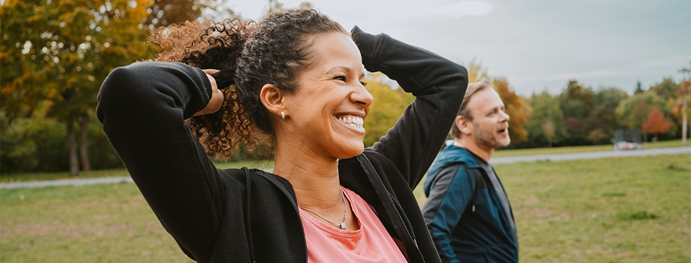 Eine Frau mit MS steht scheinbar nach dem Joggen im Park neben ihrem Trainingspartner und lächelt.
