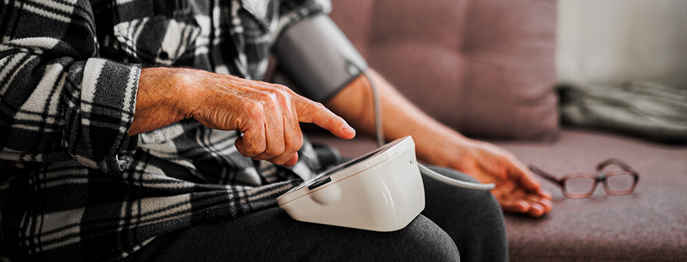 Ein älterer Mann sitzt auf der Couch und misst mit einem elektrischen Messgerät seinen Blutdruck.