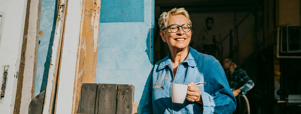 Eine Frau steht mit einer Tasse in der Hand an eine Wand gelehnt und schaut lächelnd in die Sonne, die sie anstrahlt.