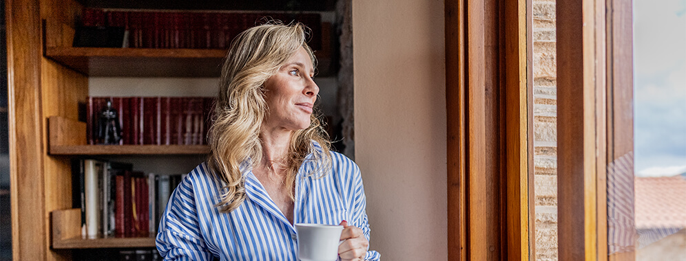 Eine Frau hält eine Tasse in der Hand und schaut aus dem Fenster.