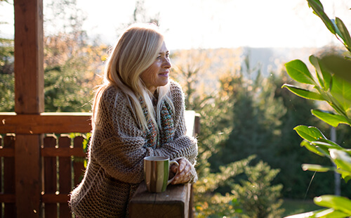 Eine Frau steht mit einer Tasse in der Hand auf einer sonnigen Terrasse.