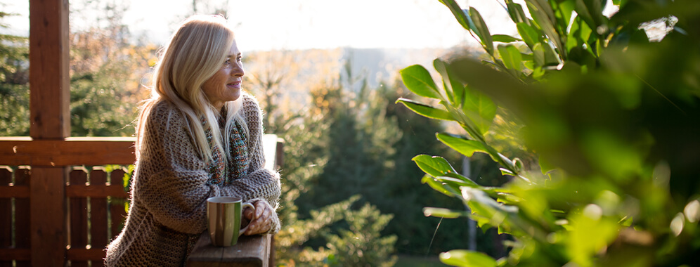 Eine Frau steht in der Sonne auf der Terrasse und hat eine Tasse in der Hand. 