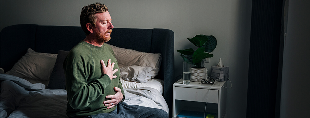 Ein Mann sitzt auf einem Bett und hält sich den scheinbar schmerzenden Oberkörper.