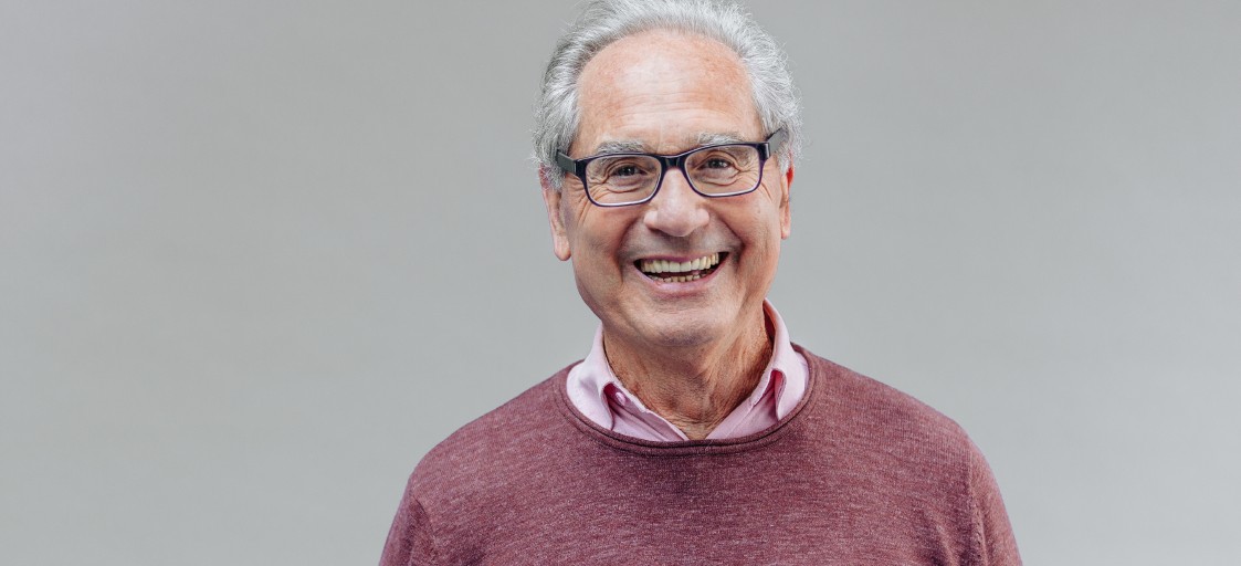 Älterer Mann mit Brille lächelt. Er trägt ein weinrotes Oberteil.