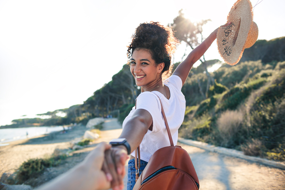 Eine Frau hält die Hand eines Mannes und lächelt in die Kamera. Sie stehen an einem sonnigen Tag am Meer.