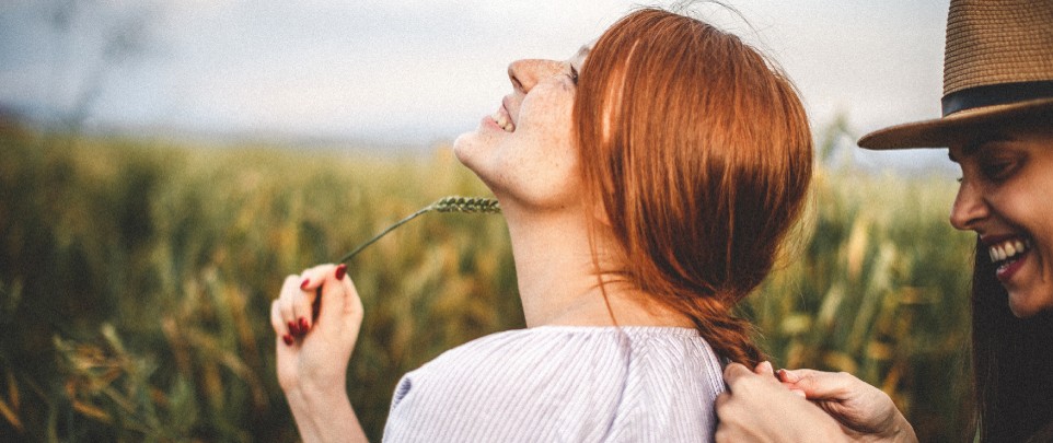 Zwei Frauen im Feld. Eine flechtet Zopf. Grüne Umgebung, sonniger Tag.
