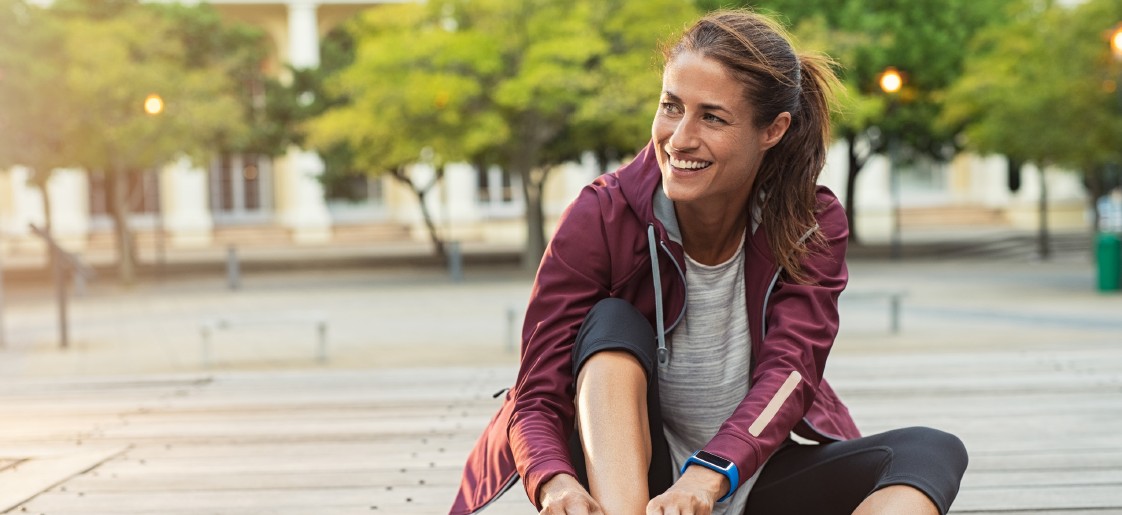 Frau sitzt draußen. Sportkleidung. Lächelt. Blaue Smartwatch.