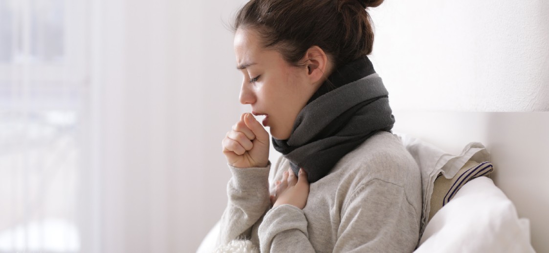 Frau hustet, bedeckt Mund und Hals mit Schal. Sie sitzt im Bett.