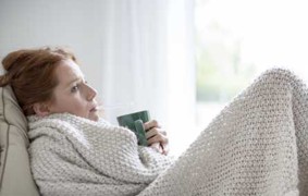 Frau liegt auf Couch, Decke, hält Tasse.