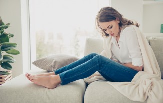 Frau sitzt auf Sofa, hält Bauch. Decke. Jeans und weißes Hemd. Fenster im Hintergrund.