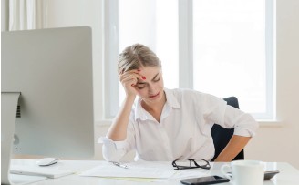 Eine Frau sitzt an einem Schreibtisch und hält sich die Hand an die Stirn. Sie wirkt gestresst.