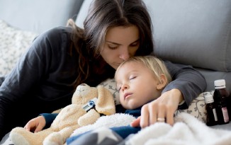 Kind schläft im Bett. Frau küsst Kind auf die Stirn. Teddybär.