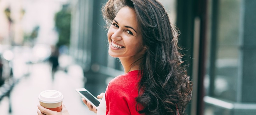 Frau mit Smartphone und Kaffeebecher. Lächelt in die Kamera.