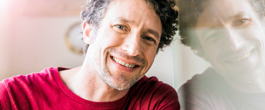 Ein Mann lächelt. Er trägt ein rotes Hemd. Spiegelung im Hintergrund.